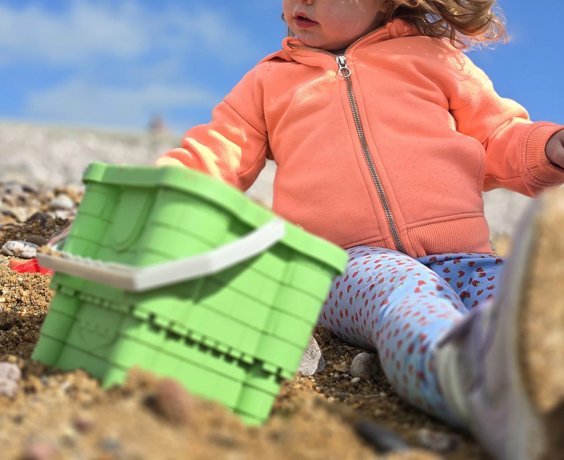 A toddler in an orange hoodie playing on the beach with a green sand bucket